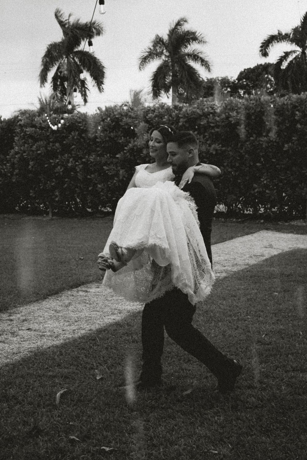 Groom carries bride in wedding dress outdoors on grass with palm trees in black and white photo.