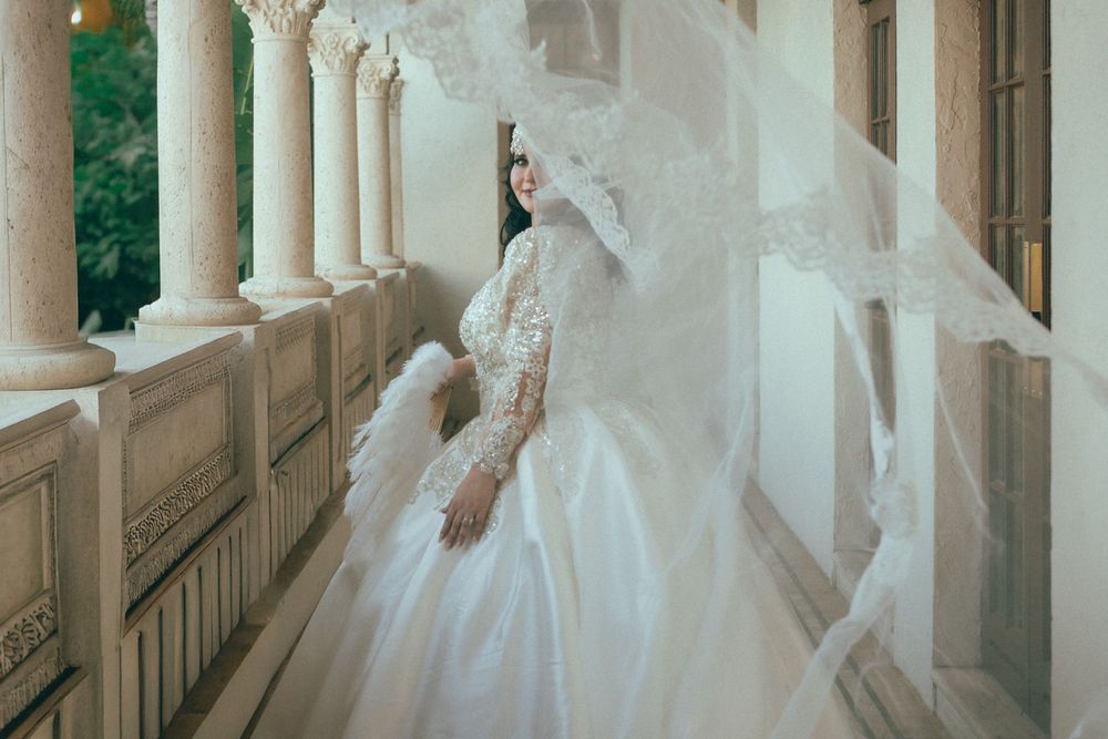 Bride in ornate ball gown with flowing veil billowing on elegant columned balcony.