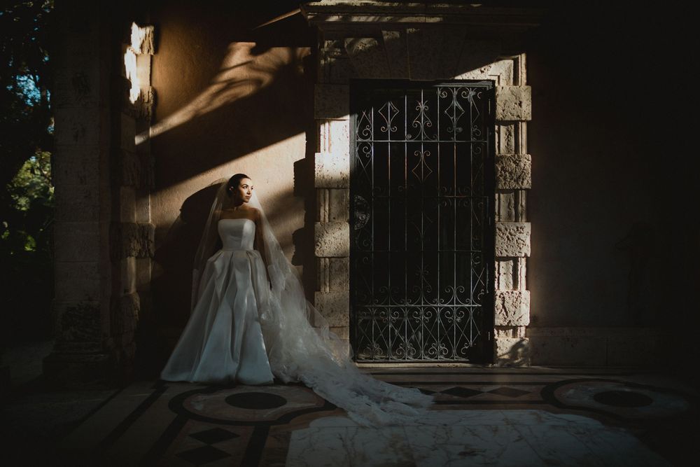 Bride in elegant white gown with long veil standing by ornate iron gate in dramatic sunlight.