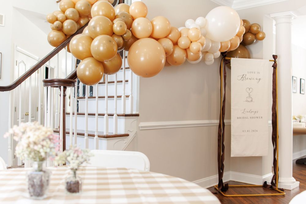 Elegant white and gold balloon garland decorates a staircase railing with framed artwork and event signage.