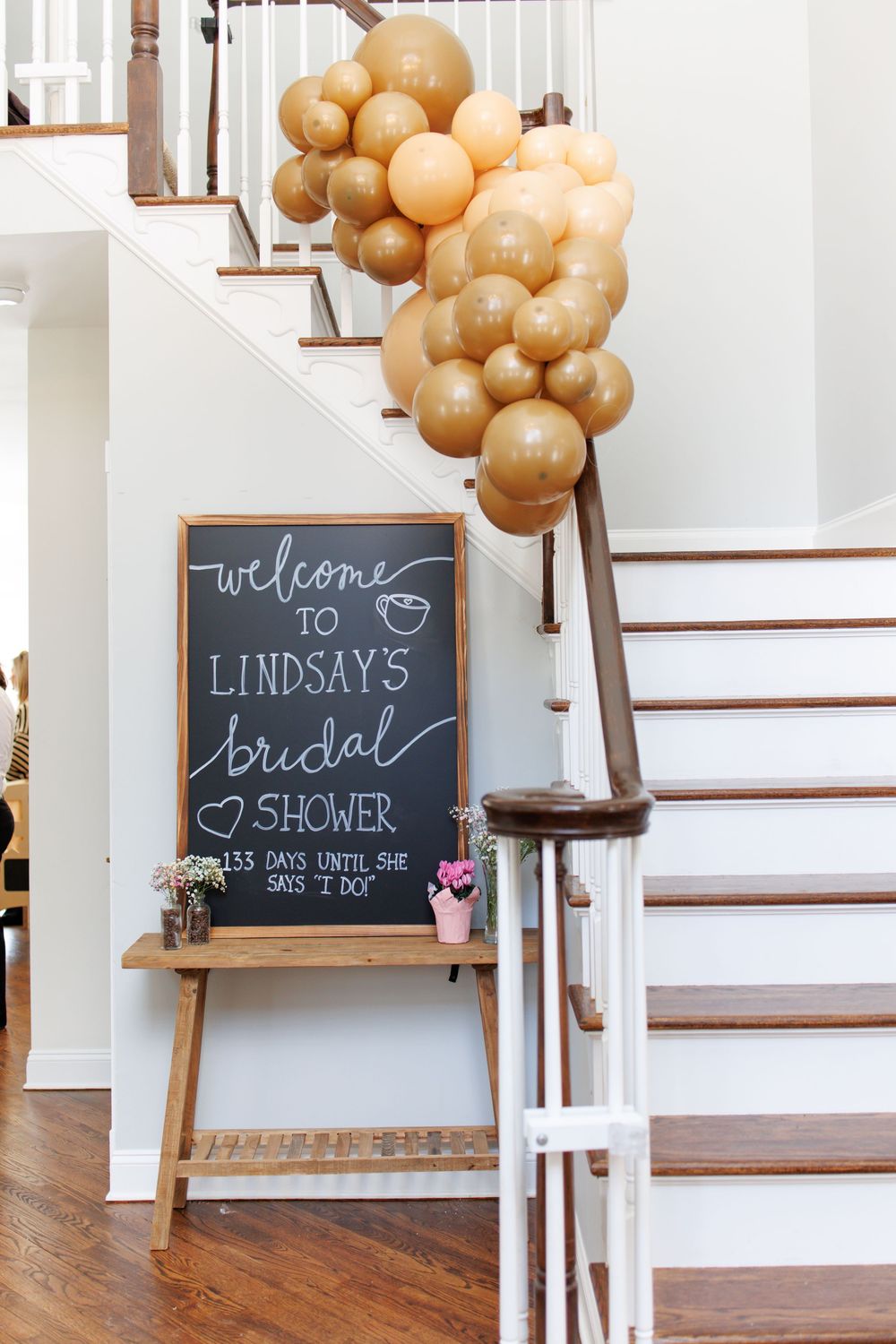 Chalkboard welcome sign and gold balloon decorations set up near wooden staircase for bridal shower celebration.