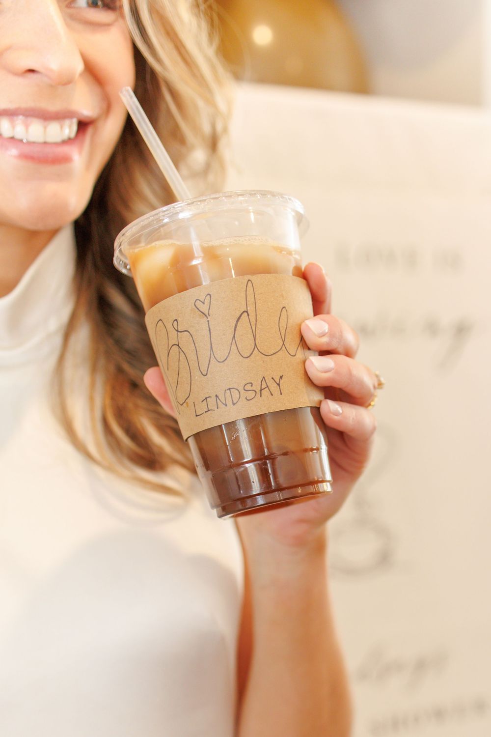 Hand holding an iced coffee drink in a clear plastic cup with writing on it against a light background.