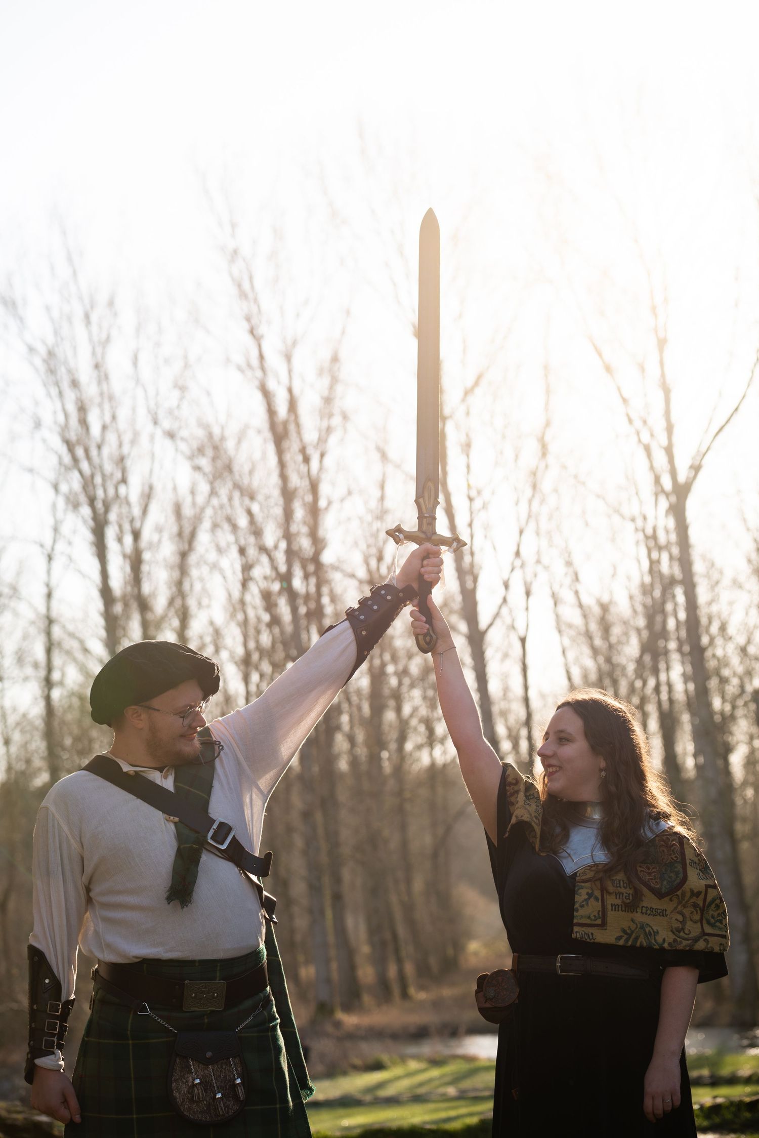 Two people in medieval costumes raise their swords together in a wooded outdoor setting at sunset.