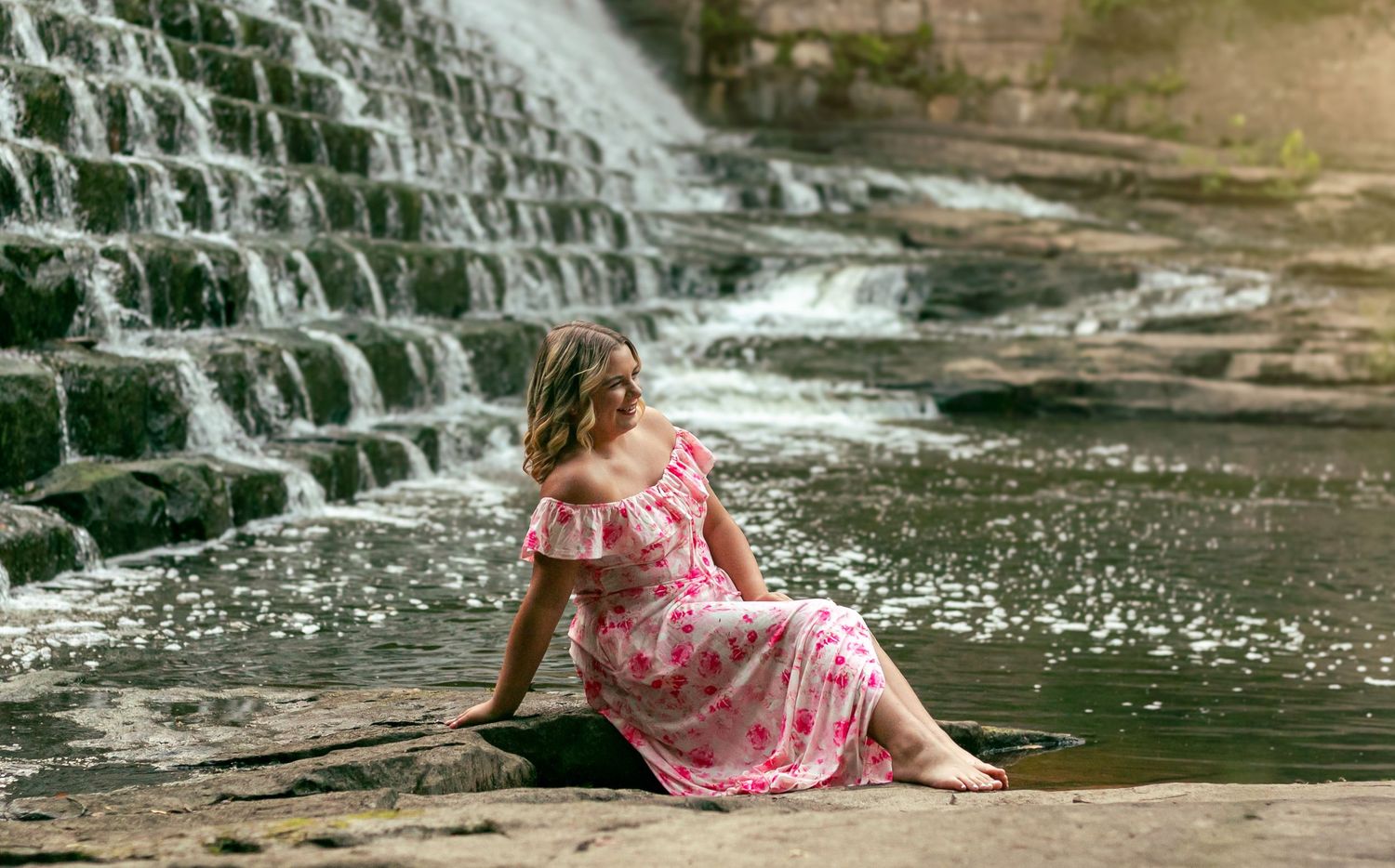 A woman in a pink floral dress sits peacefully by a cascading waterfall on a sunny summer day.milll creek park Youngstown Ohio