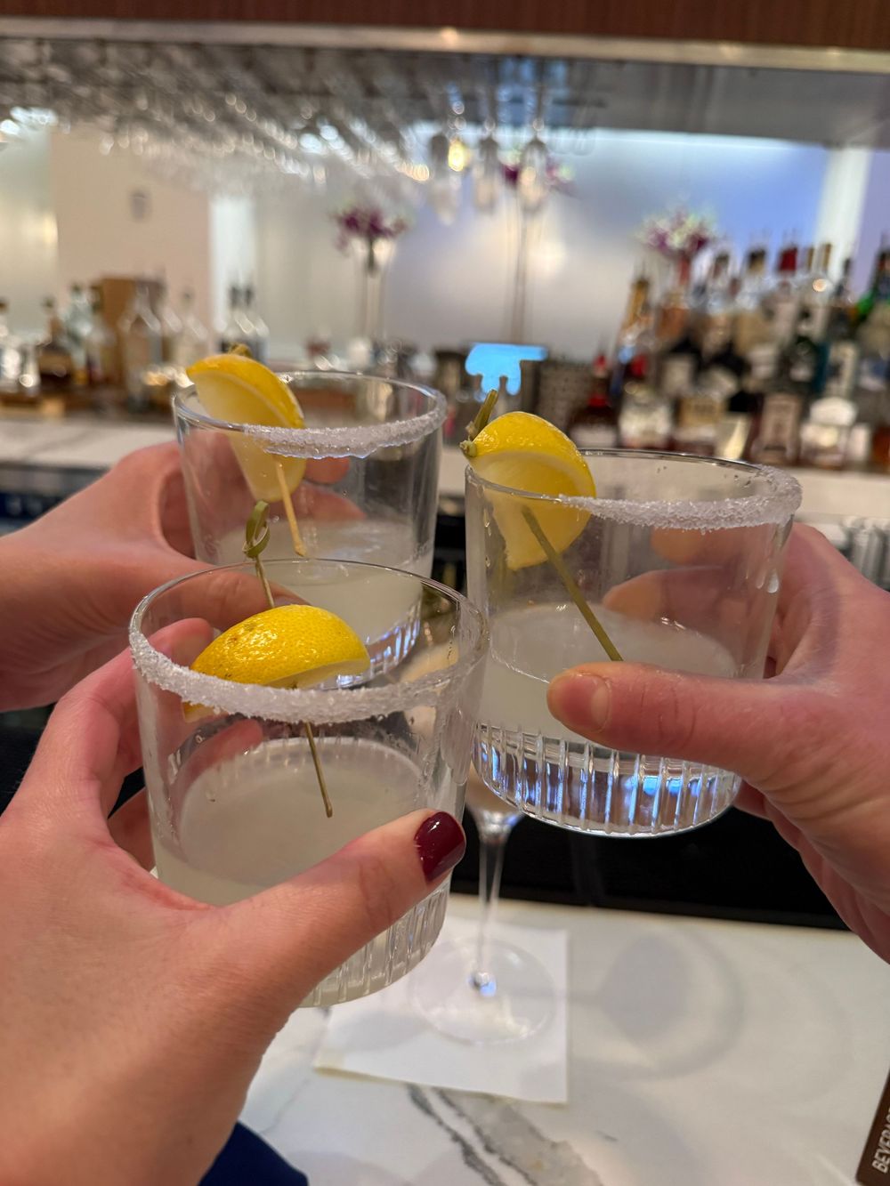 Multiple glasses of cocktails with lemon garnishes being toasted in a cheers gesture at a restaurant or bar.