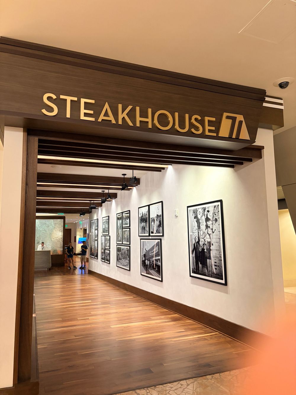 Long hallway gallery space with wooden floors, black and white photographs on white walls, and a steakhouse sign visible.