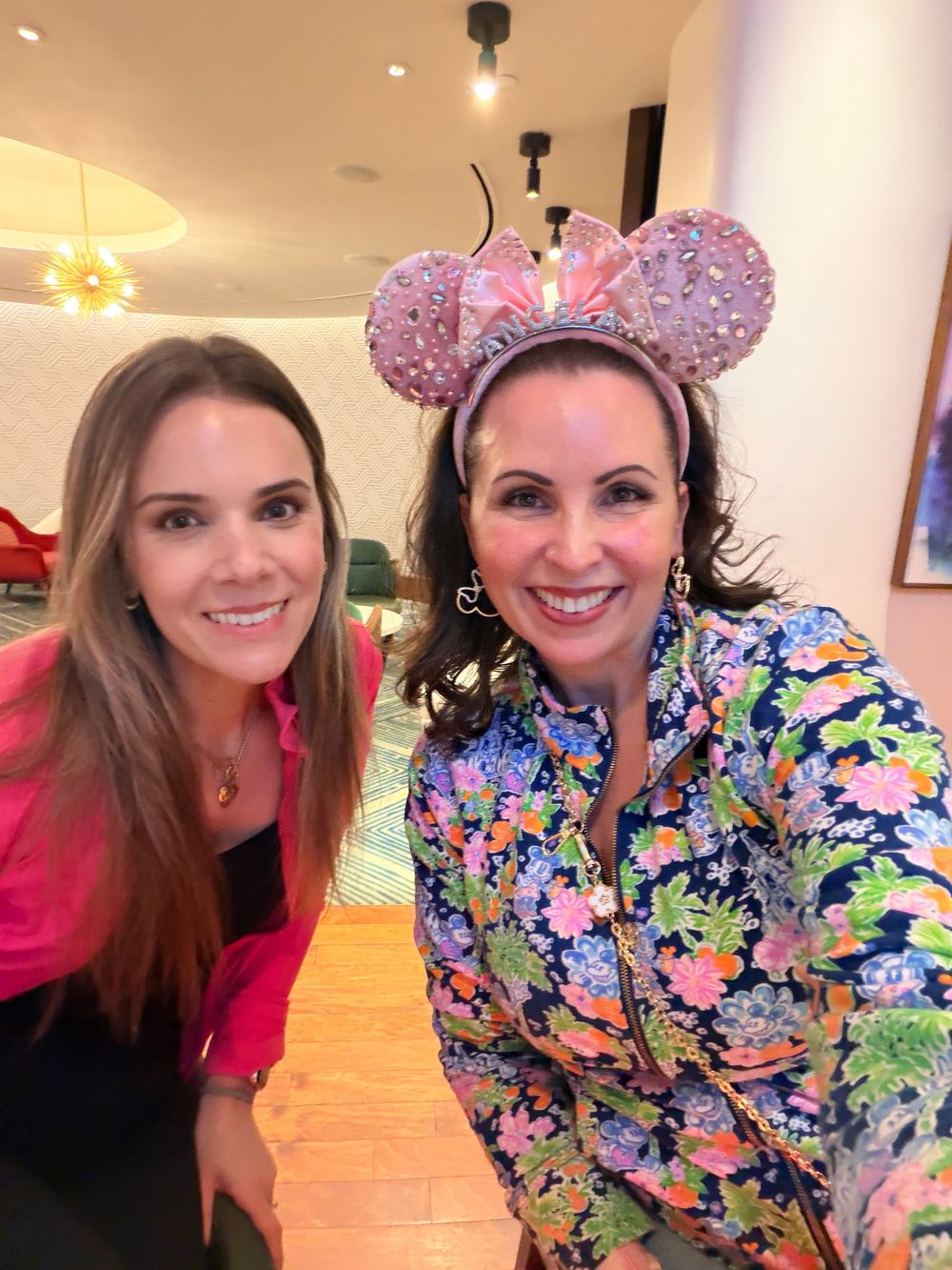 Two people pose together wearing colorful floral outfits and sparkly Minnie Mouse ears in an indoor setting.
