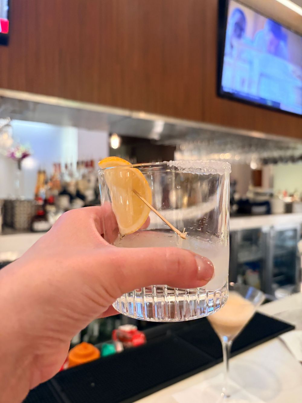 Four hands holding up cocktail glasses with clear drinks in a toasting gesture at what appears to be a bar setting.