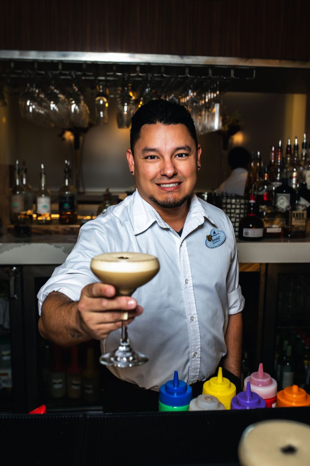 A bartender in a white shirt presents a series of colorful cocktails in martini glasses at a dimly lit bar counter.