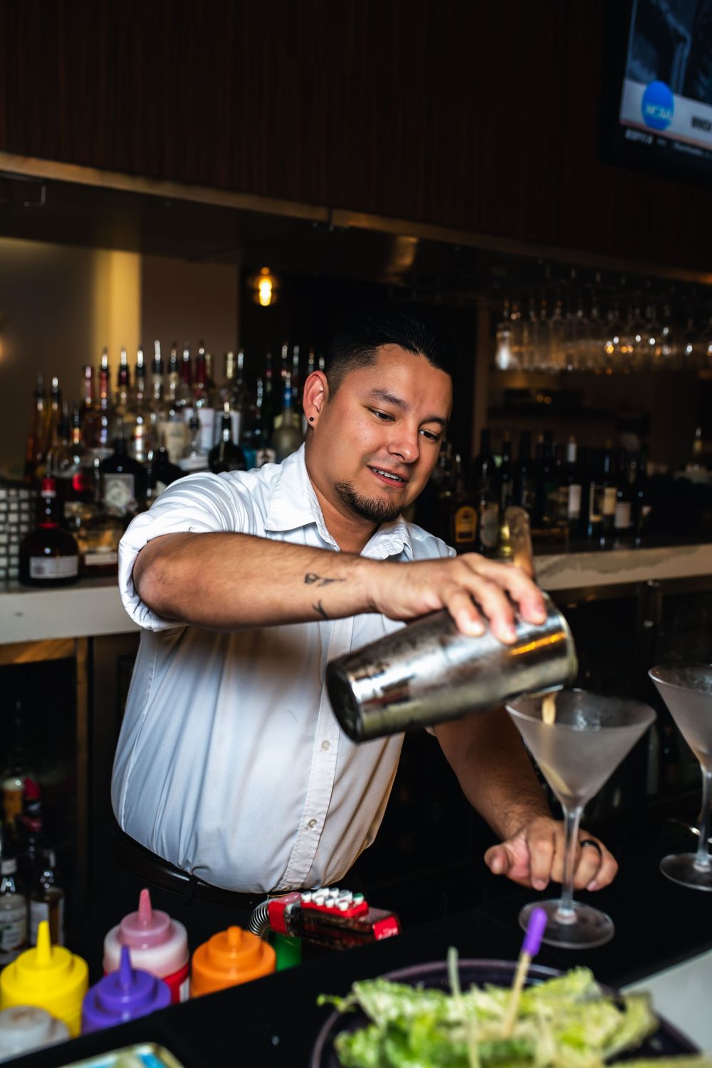 A bartender in a white shirt pours cocktails into martini glasses at a well-stocked bar.