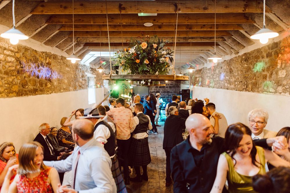 Wedding guests dance and celebrate in a rustic stone-walled venue with warm lighting and exposed beams.
