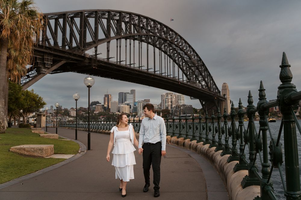A couple walks along the waterfront path beneath the iconic Sydney Harbour Bridge on a cloudy day during an engagement photoshoot