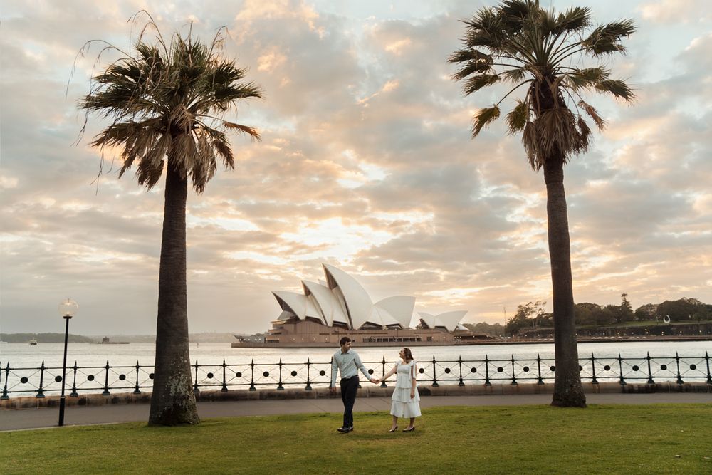 Two palm trees frame the Sydney Opera House at sunrise by the harbour where a couple walks in the middle during their engagement photoshoot