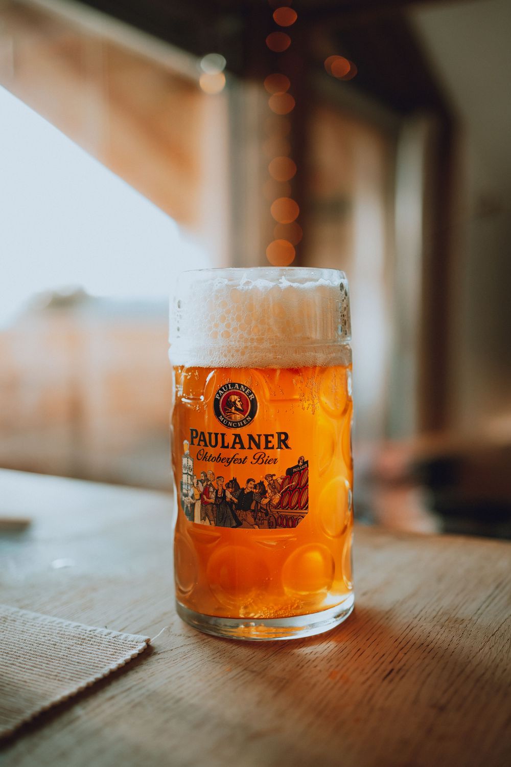 Three glasses of golden Paulaner beer sitting on a wooden table with bokeh lights in the background.
