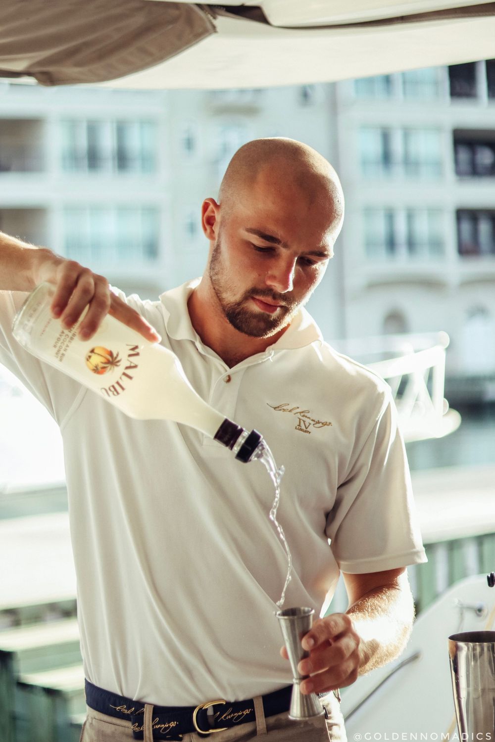 A mixologist in a white polo shirt pouring a drink on a luxury catamaran with city buildings in the background.