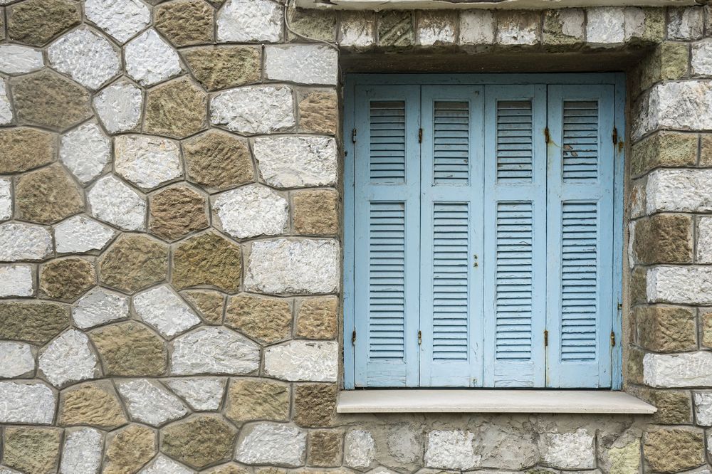 Light blue wooden shuttered window on a rustic stone wall with irregular white and beige stone pattern.