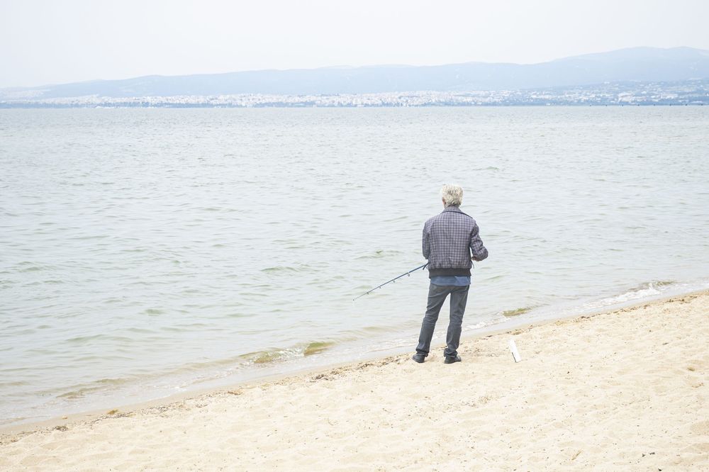 A person stands alone on a peaceful beach shoreline overlooking calm waters and distant mountains.