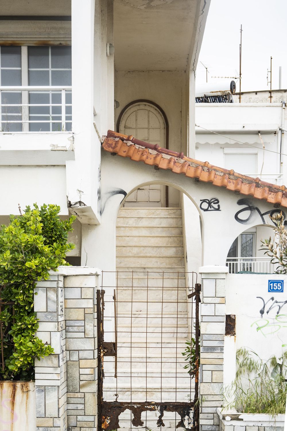 White building entrance with arched doorway, terracotta roof tiles, and metal gate surrounded by greenery.