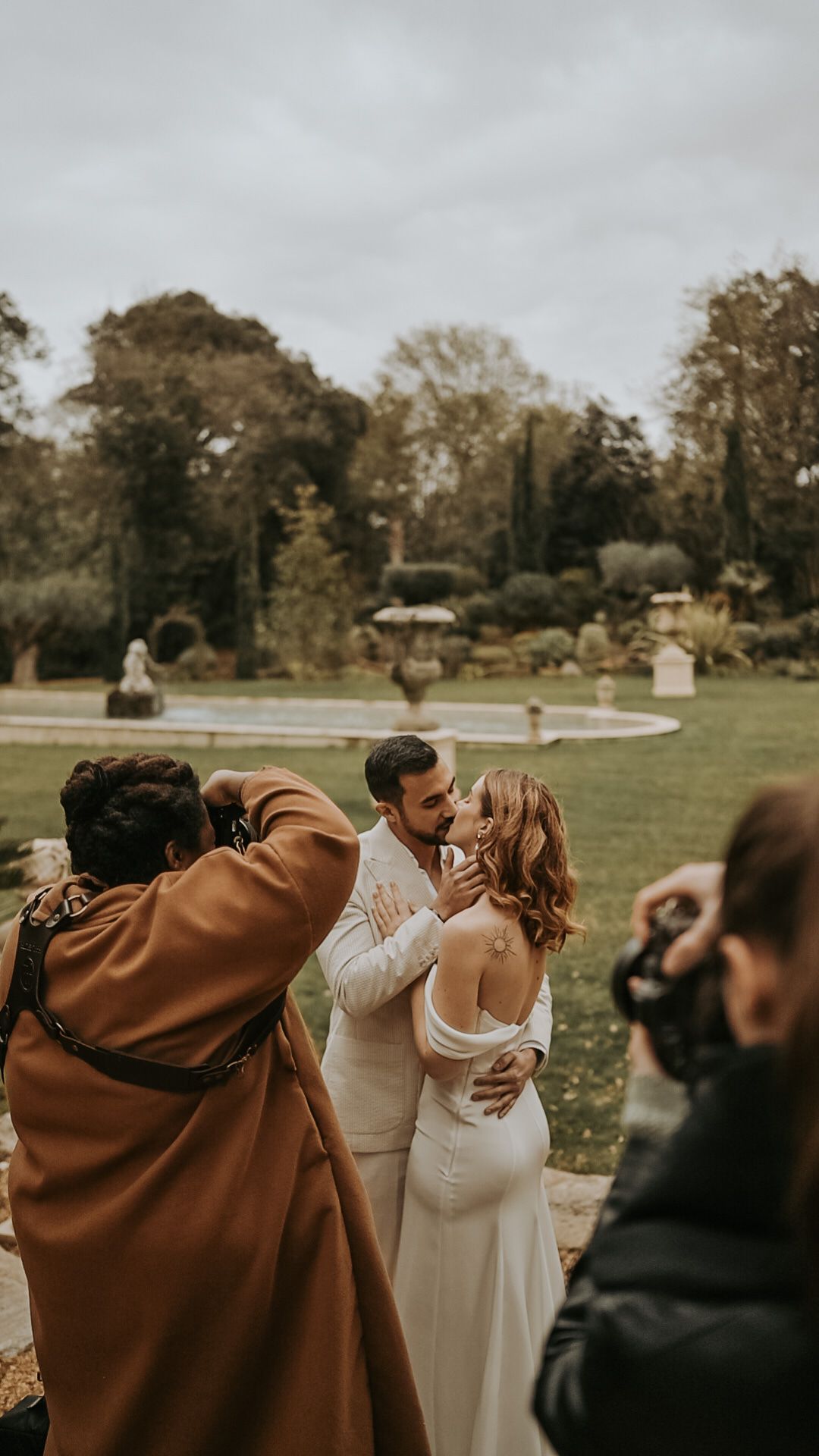 Wedding guests wrap themselves in cozy brown blankets while standing together in an outdoor garden setting.