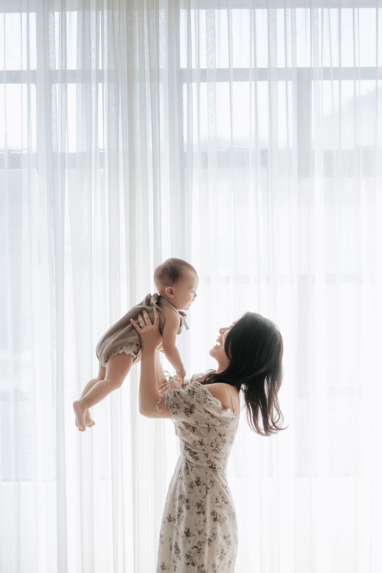 A mother in a floral dress lifts her smiling baby in the air near sheer white curtains in a bright room.一位穿著碎花洋裝的母親將她笑容燦爛的寶寶舉到空中，站在明亮的房間裡