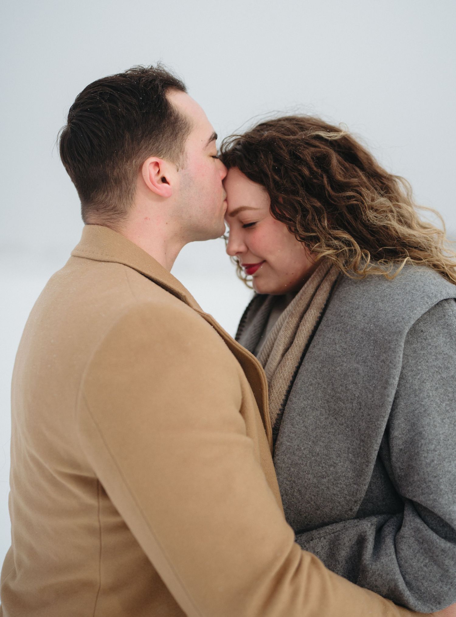 A romantic winter scene shows a couple in tan and grey coats sharing an intimate moment against a white background.