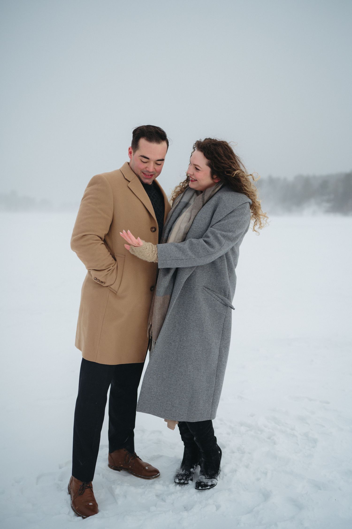 A couple playfully embraces in a snowy winter landscape wearing tan and grey coats.