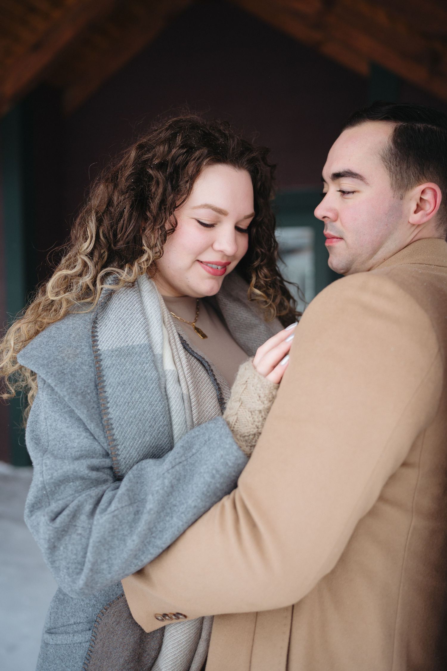 A couple in winter clothing share an intimate embrace, with the woman wearing a gray sweater and the man in a tan coat.