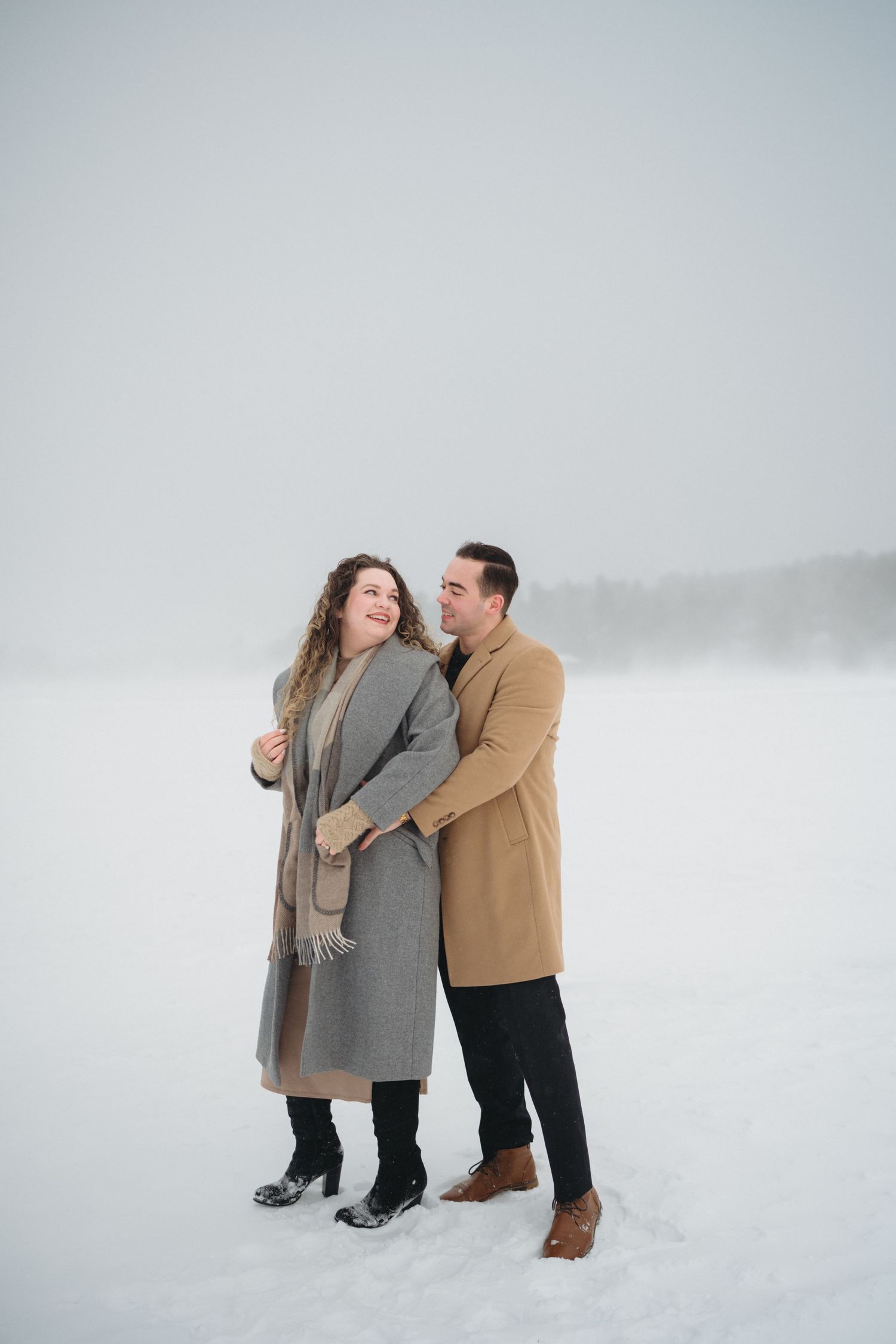 A couple shares a playful moment in the snow wearing winter coats and dark pants.