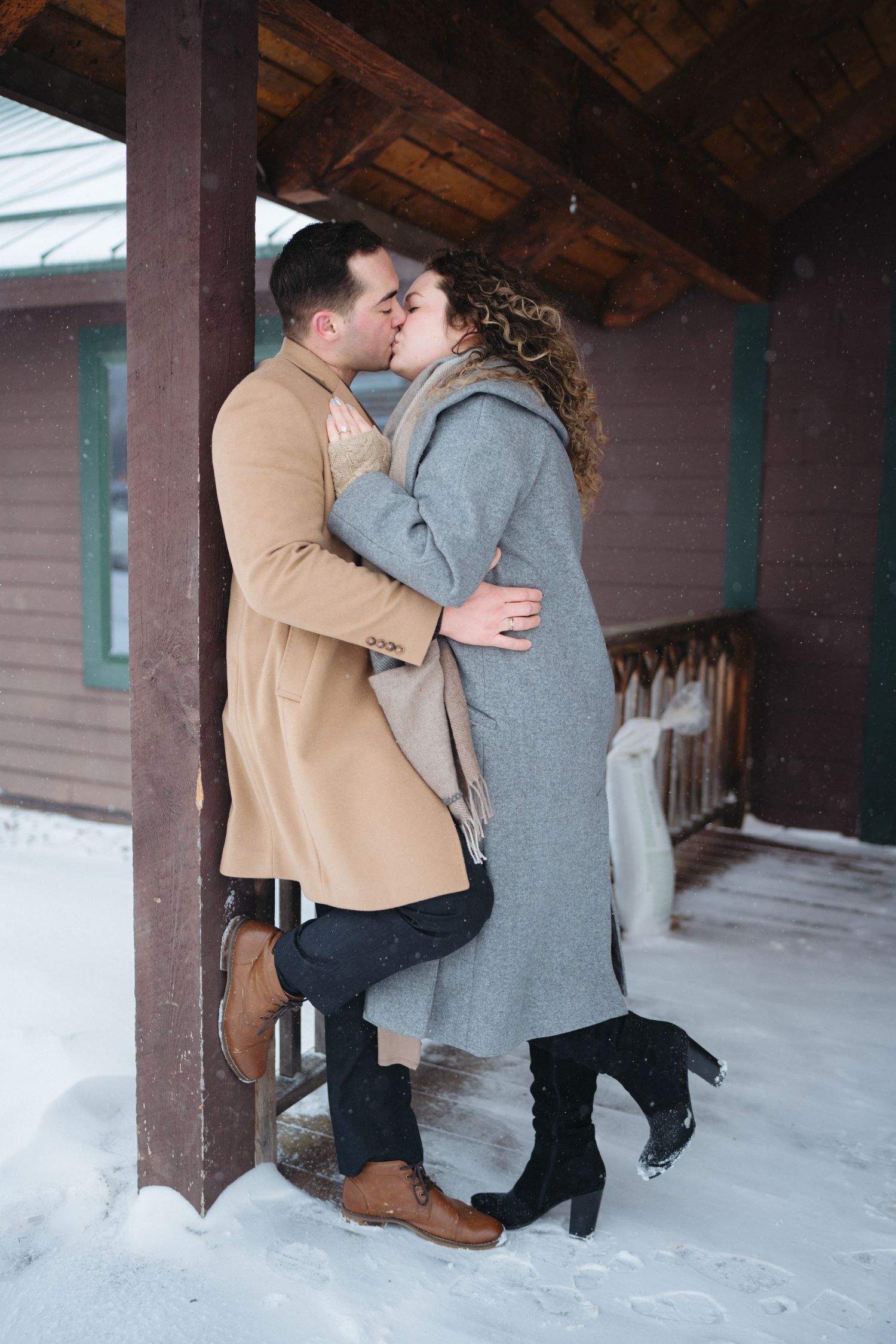 A couple shares a tender moment in the snow under a rustic wooden porch during winter.