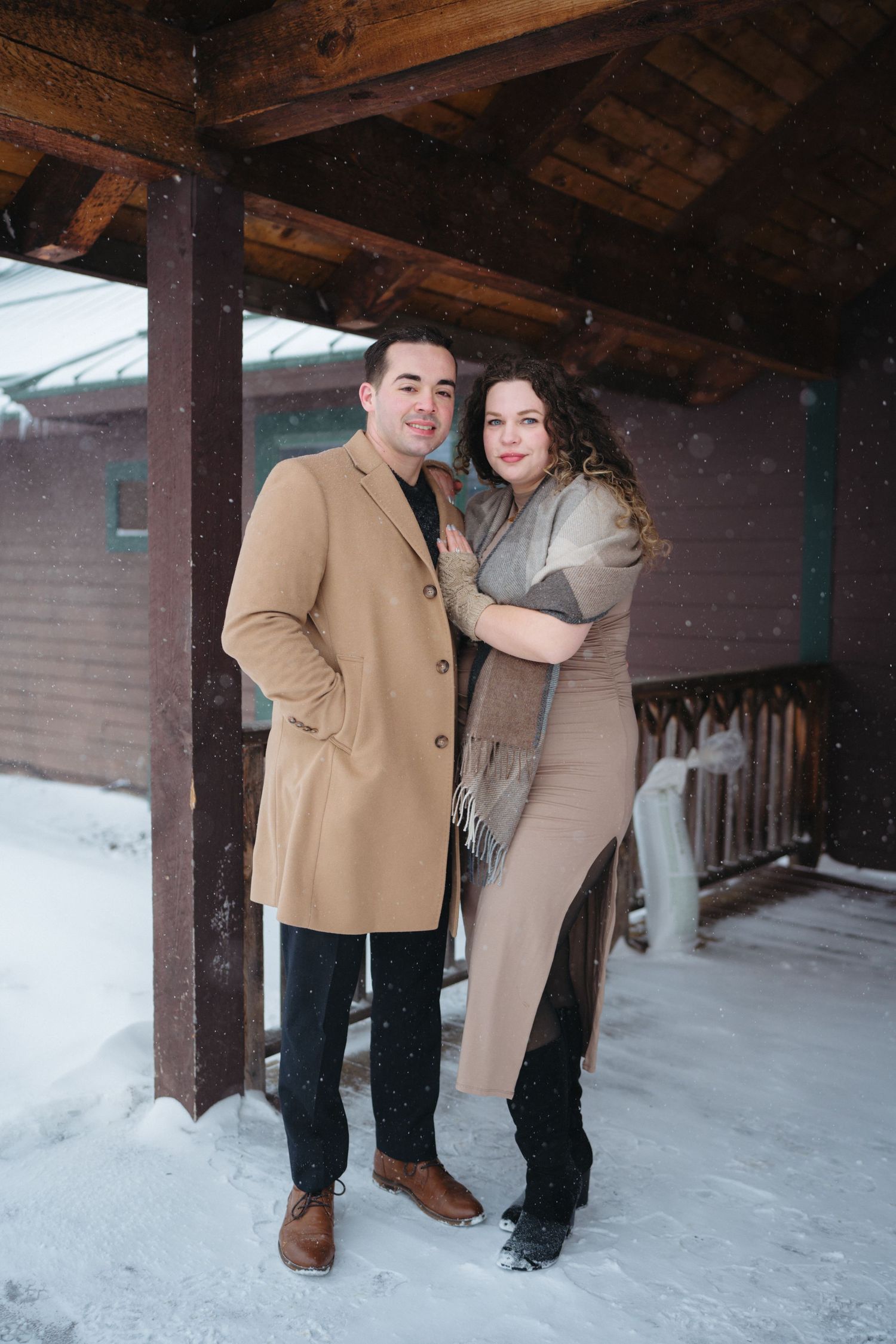 A couple poses together on a snowy porch wearing coordinating camel and beige winter outfits.