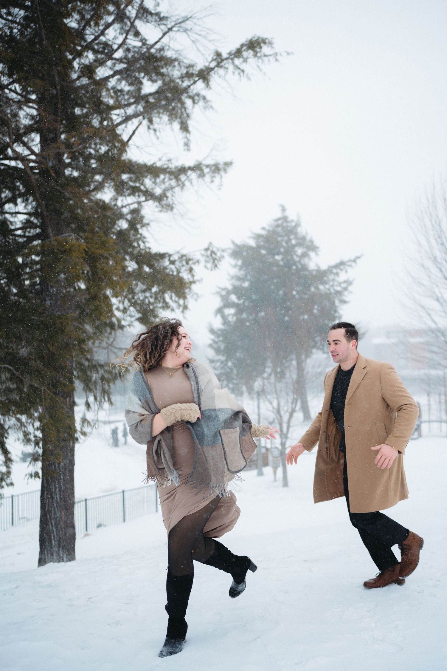 A playful couple frolics in a snowy winter landscape with evergreen trees and fog in the background.