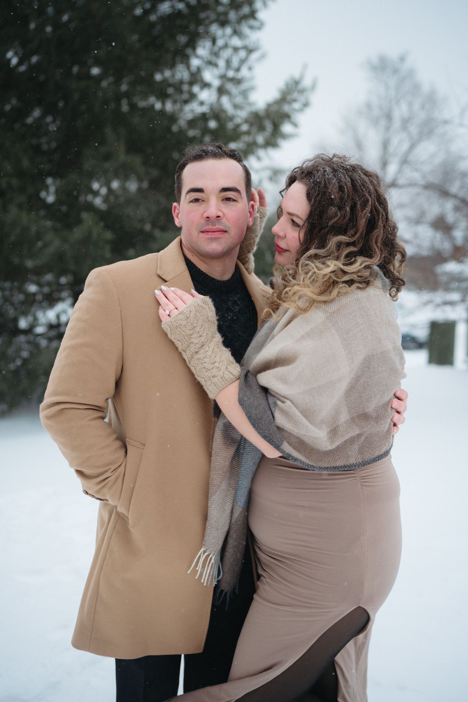 A romantic couple poses playfully in a snowy winter landscape, wearing cozy coats and scarves.