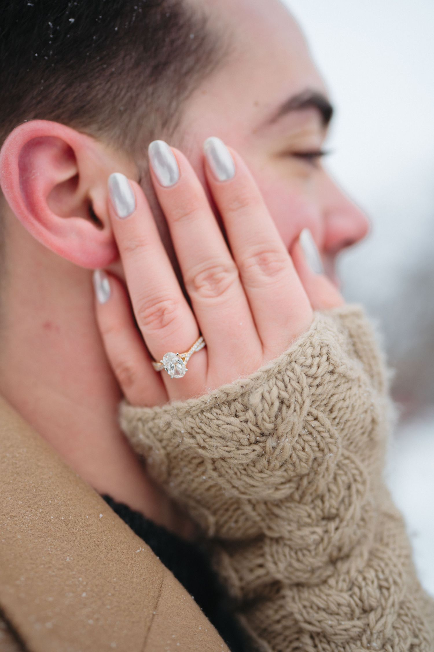 Close up of hands wearing a knit sweater and engagement ring against a snowy winter background.