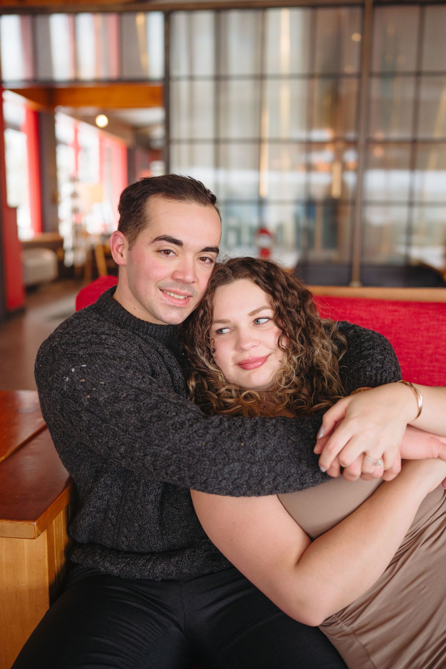 A cozy indoor portrait of a couple embracing on red furniture wearing dark winter sweaters.