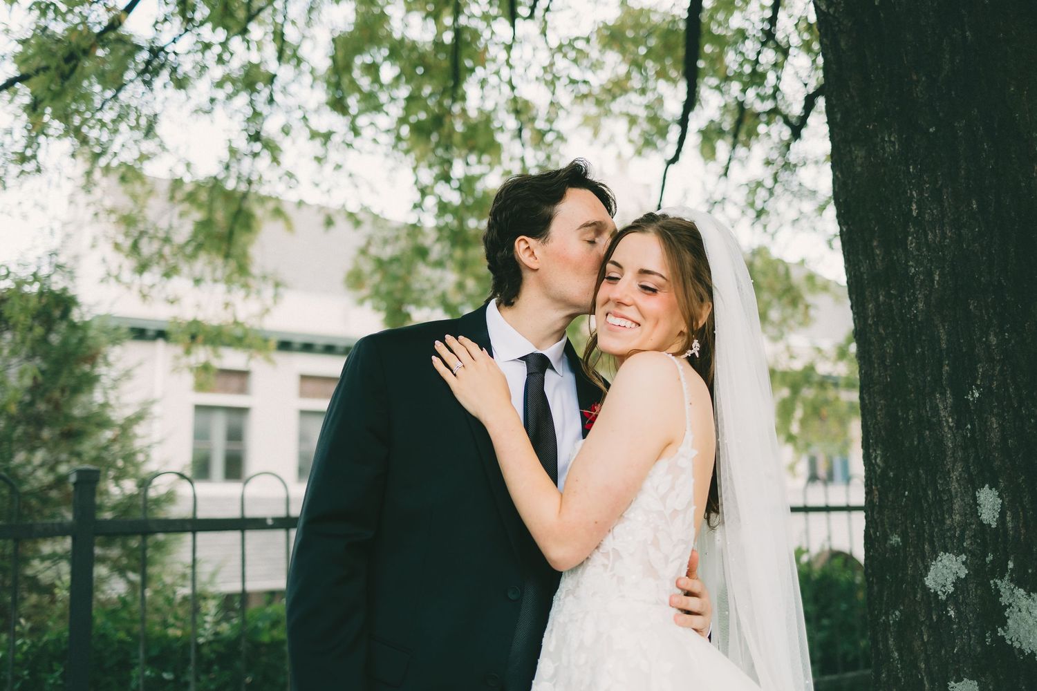 A couple shares an intimate moment while embracing outdoors under tall trees.