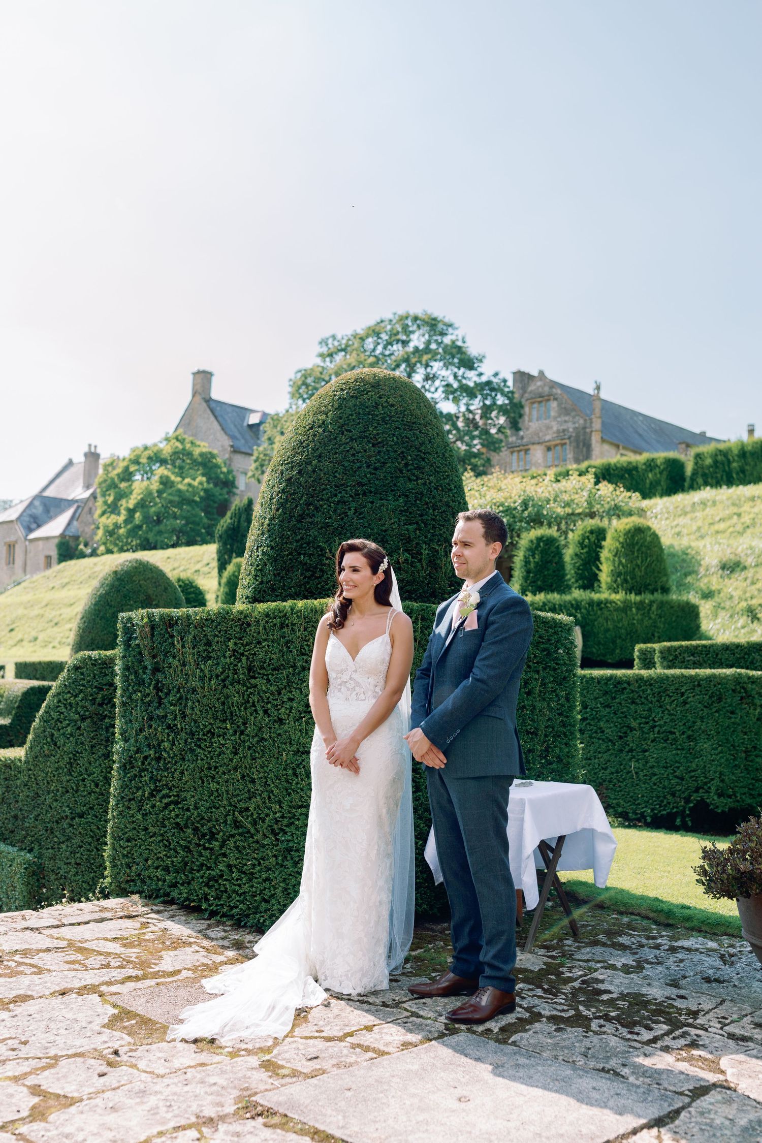A couple stands together at their wedding ceremony in a garden with beautifully manicured topiary hedges in the background.