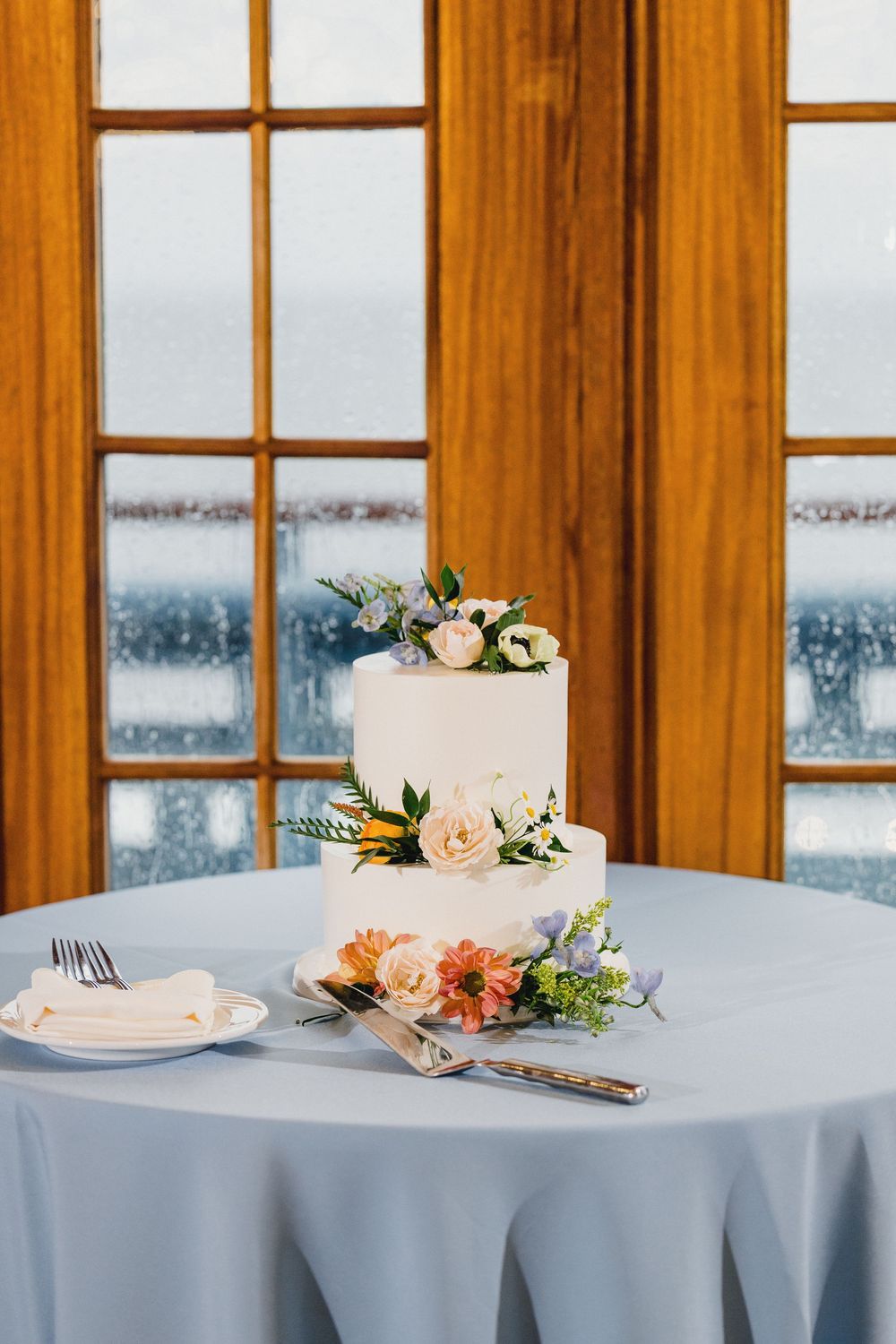 Elegant two-tier white wedding cake with peach and blue floral decorations on light blue tablecloth by frosted windows.