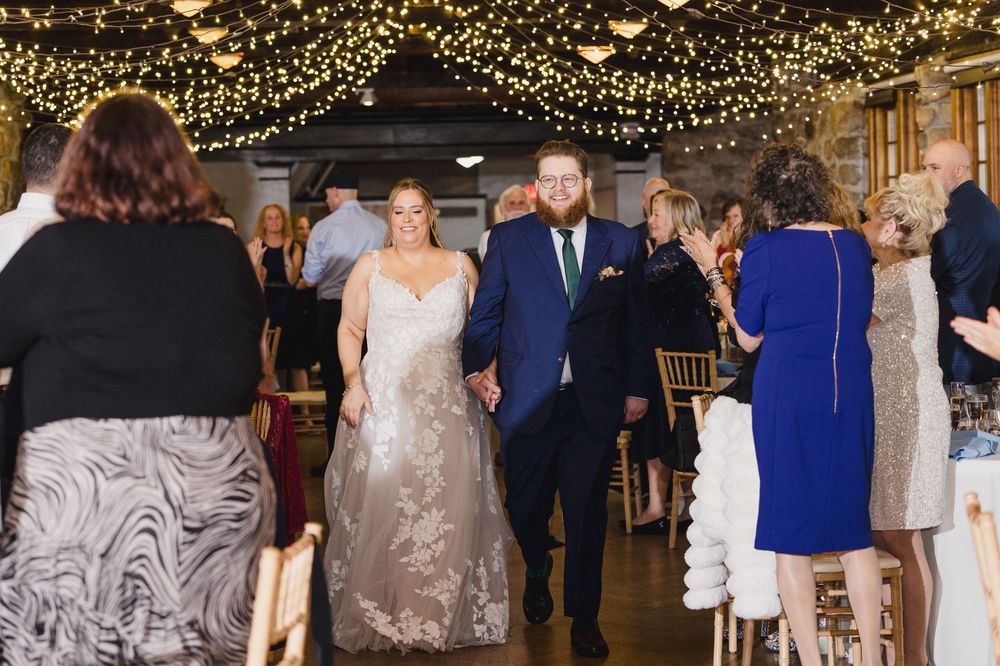 Wedding reception in a rustic venue decorated with twinkling string lights overhead as couple walks down the aisle.