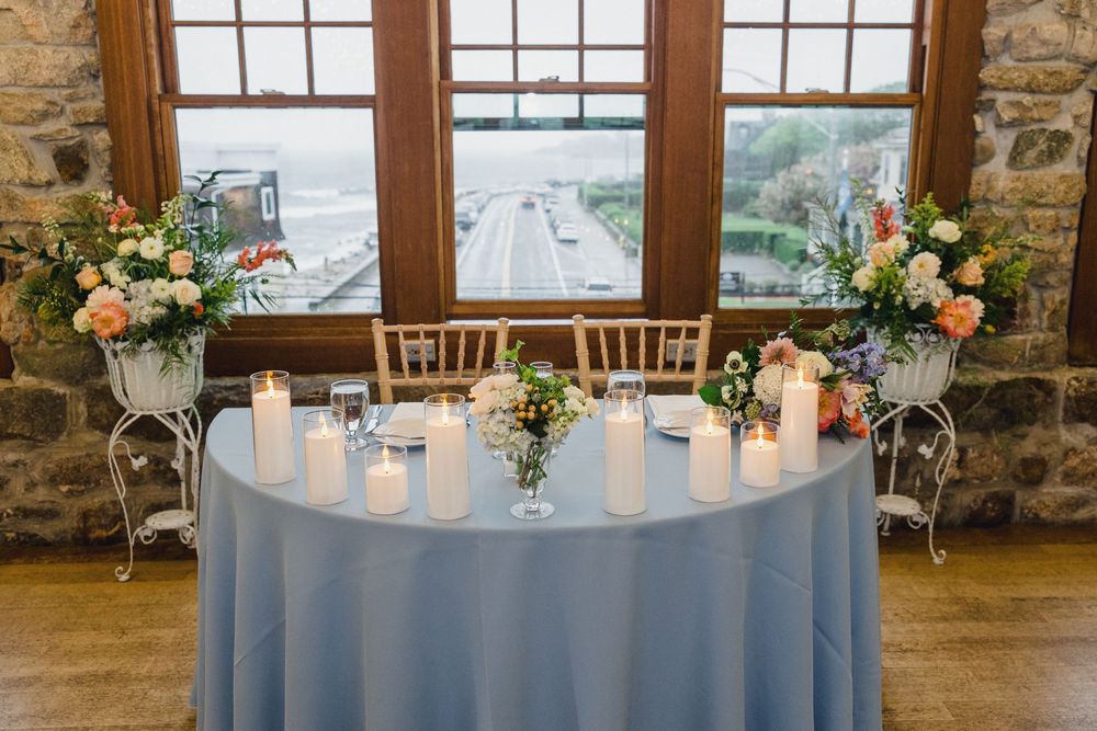 Elegant wedding reception table with light blue tablecloth and floral centerpieces next to stone wall windows.