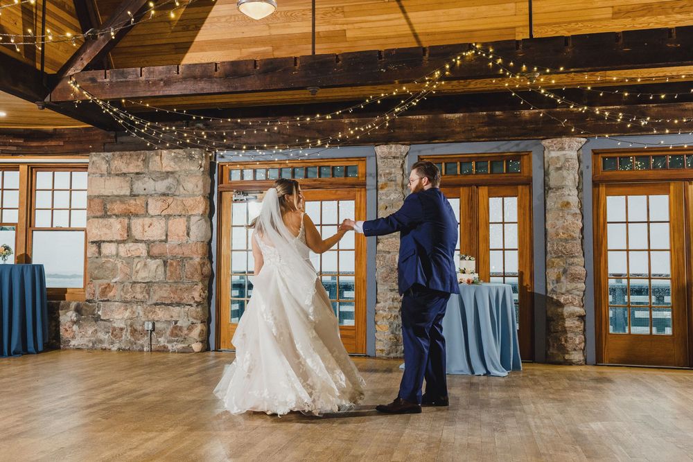 Couple dancing in rustic venue with stone walls, wooden ceiling, and string lights.