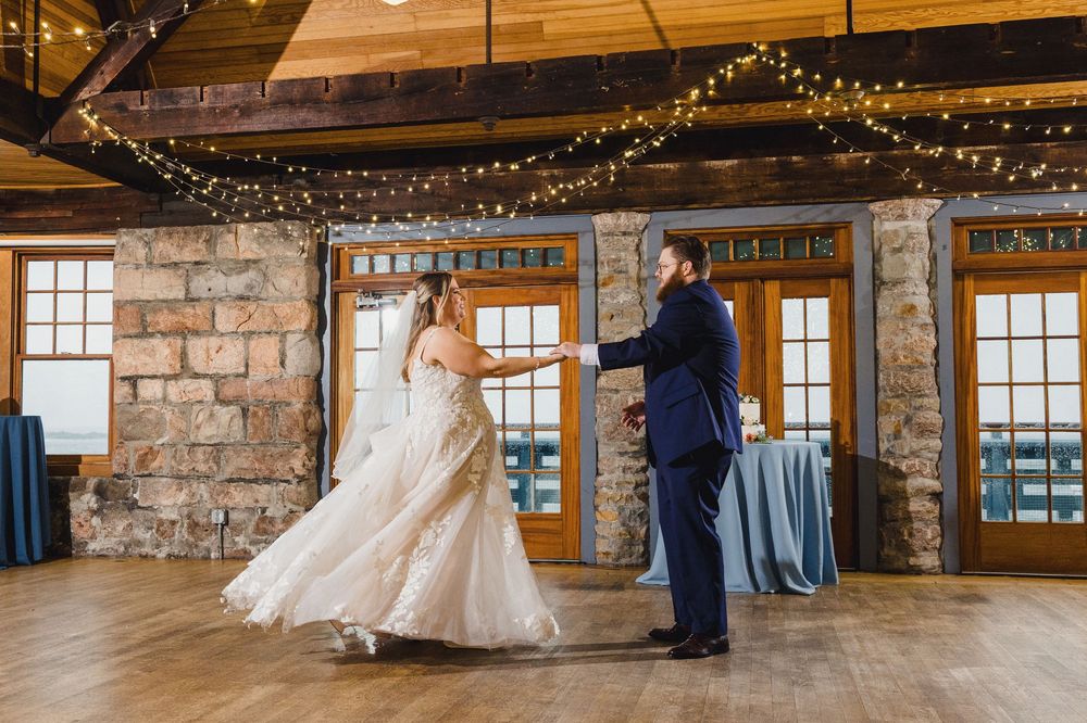 A wedding couple dances in a rustic stone-walled venue decorated with twinkling string lights overhead.