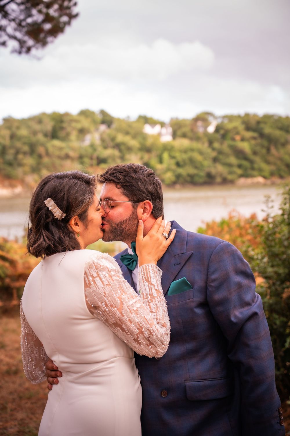 A bride and groom share a romantic kiss outdoors, with a scenic river and green hills in the background.