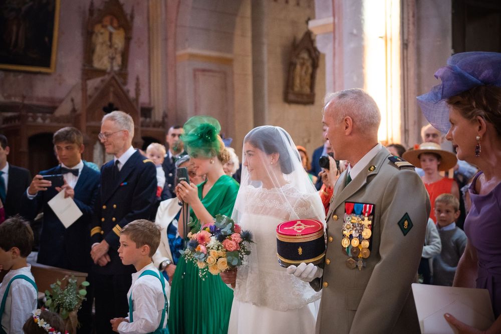 Bride in white veil holds bouquet beside military officer with medals in ornate church wedding ceremony.
