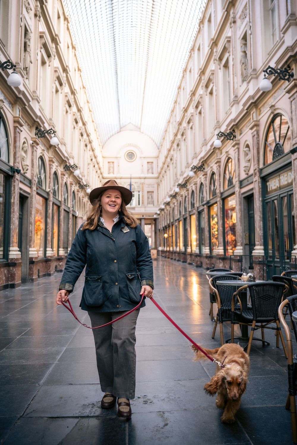 Woman in hat walks golden cocker spaniel on red leash through elegant covered shopping gallery.