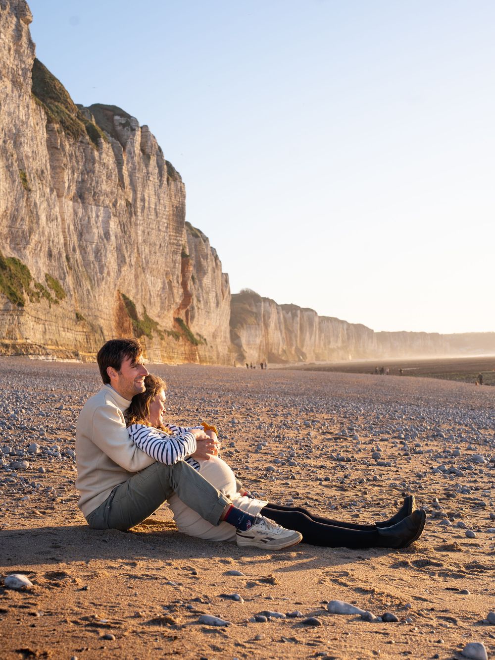 A man and child sit together on a pebble beach beside towering white chalk cliffs at golden hour.