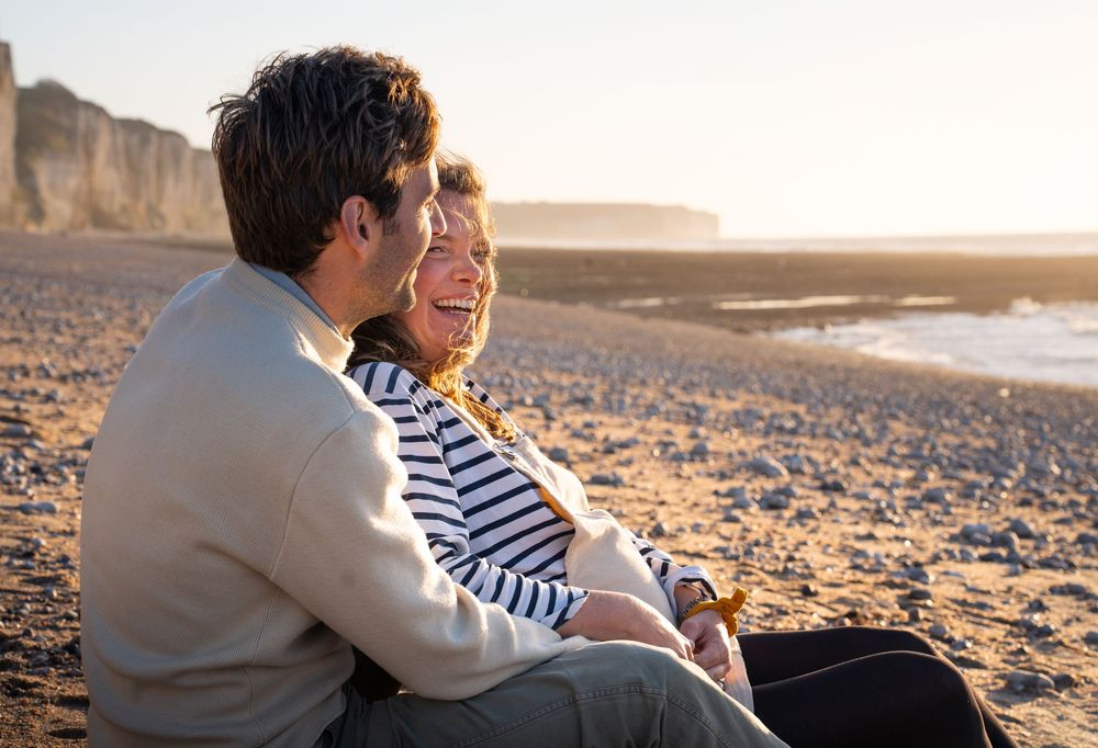 Happy couple sitting together on a sunny beach, smiling and relaxing near coastal cliffs at golden hour.