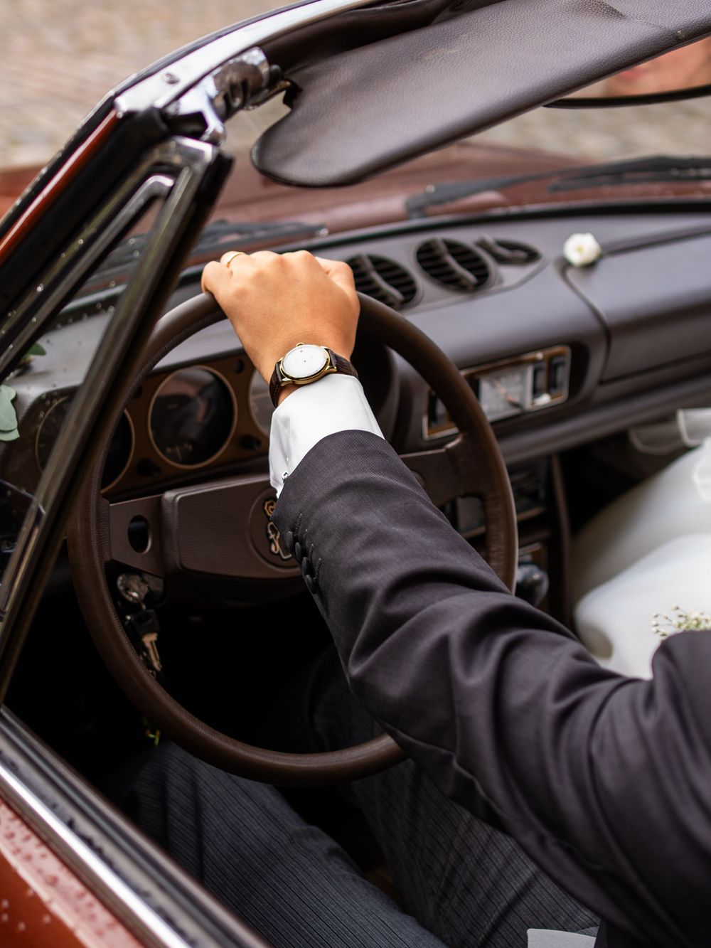 Man in suit wearing a gold watch driving a vintage convertible car with brown leather steering wheel.