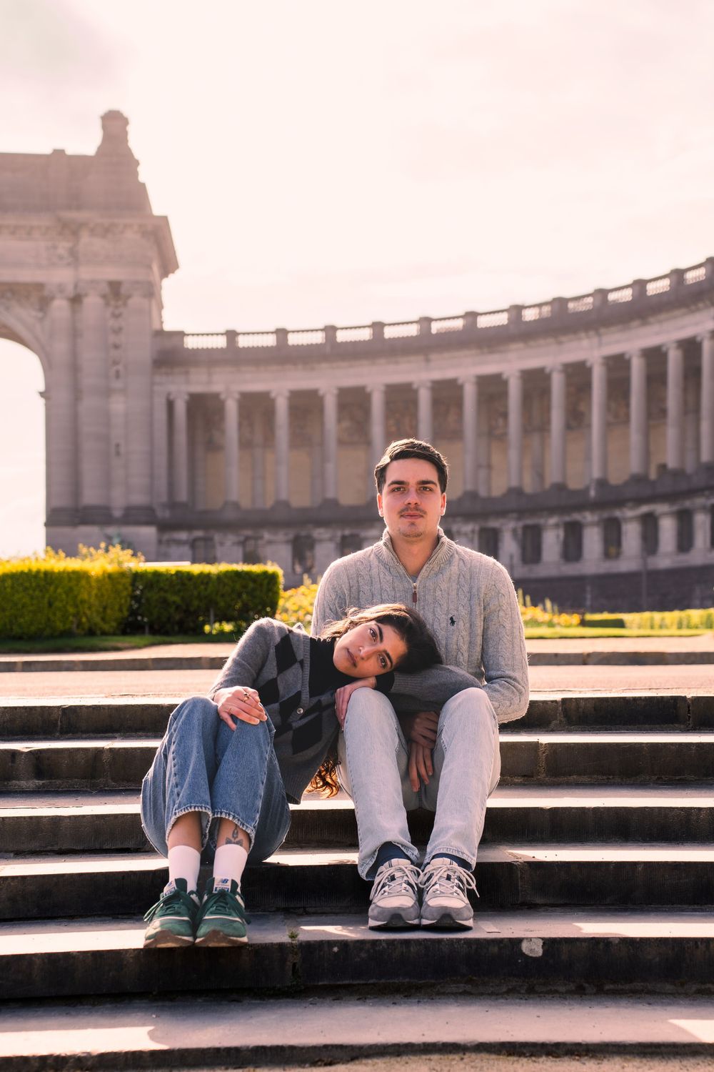 Couple sitting on steps in front of a grand neoclassical monument with colonnade, sharing a tender moment outdoors.