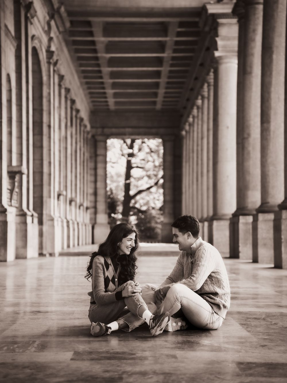 Couple sitting on marble floor of elegant columned hallway, sharing an intimate moment in sepia-toned romantic photo.