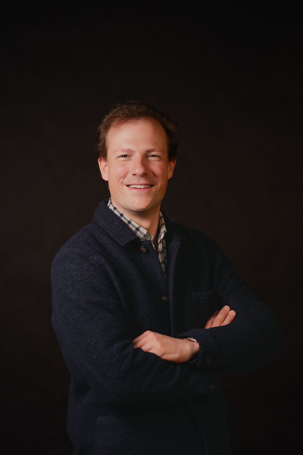 Smiling man with crossed arms wearing a navy sweater over plaid shirt, posing against a dark studio background.